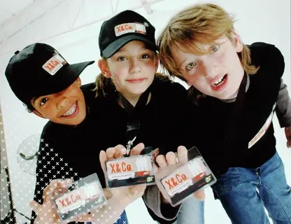 Drei Kinder mit schwarzen Cappies blicken in die Kamera u nd halten einen Ausweis mit dem Schriftzung XundCo. hoch.