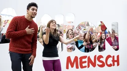 Andreas Bourani und Lisa tanzen vor dem Logo der Aktion Mensch. Im Hintergrund sind lachende Menschen zu sehen.