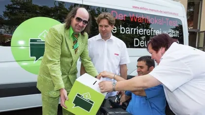 Guildo Horn, Michael Wahl, Petra Groß und ein Mann im Rollstuhl halten gemeinsam eine Wahlurne aus Karton in den Händen.