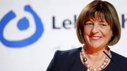 Eine Frau, Ende 60, mit braunen Haaren im Pagenkopfschnitt, geschminkten Lippen einer Kette mit dicken Kugeln und blauem Kostüm lächelt leicht an der Kamera vorbei. Im Hintergrund etwas verschwommen das Logo der Lebenshilfe.