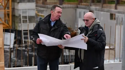 Zwei Männer stehen mit einem Bauplan in der Hand  vor einer Baustelle, auf der ein Haus gebaut wird.