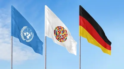 Drei Flaggen an Mast vor blauem Himmel: Eine mit dem Logo der UN, eine mit vielen bunten Punkten im Kreis, die Inklusion symbolisieren sollen und die deutsche Fahne