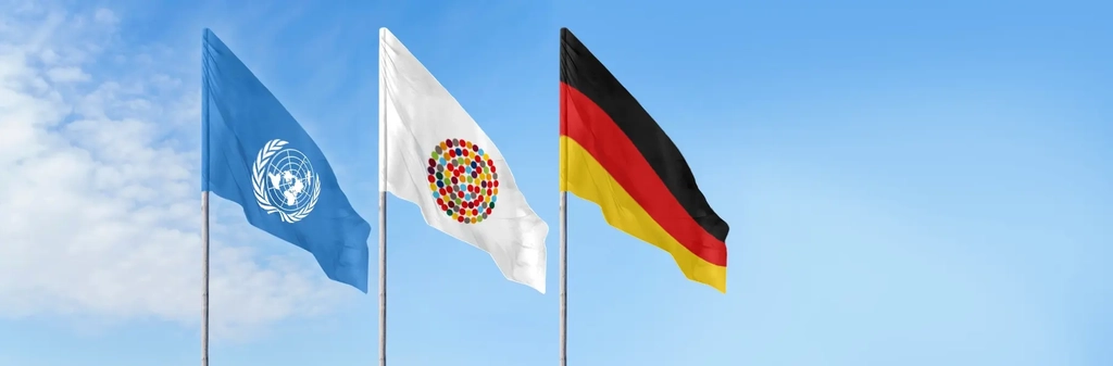 Drei Flaggen an Mast vor blauem Himmel: Eine mit dem Logo der UN, eine mit vielen bunten Punkten im Kreis, die Inklusion symbolisieren sollen und die deutsche Fahne
