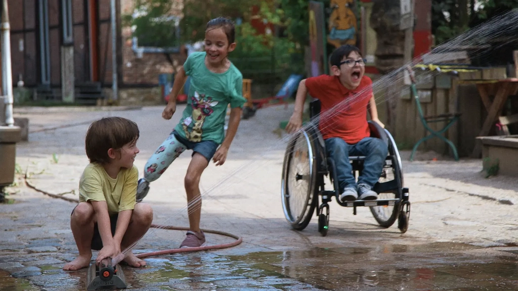 Drei Kinder spielen gemeinsam auf einem Bauernhof. Sie verspritzen Wasser aus einem Gartenschlauch und lachen ausgelassen. Eines der Kinder benutzt einen Rollstuhl, ein anderes trägt eine Prothese am rechten Bein.