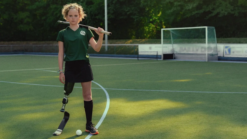 Annika, ein Mädchen mit lockigem Haar und Beinprothese steht auf einem Hockeyplatz. Zwischen ihren Beinen liegt der Ball, ihren Schläger hat sie lässig über die Schultern gelegt.