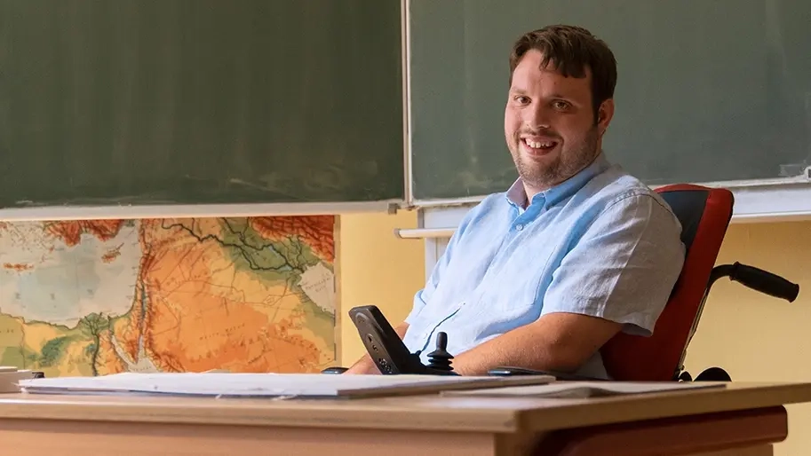 Ein junger Mann in einem Elektrorollstuhl sitzt hinter einem Lehrerpult in einem Klassenzimmer. Hinter ihm ist eine große Landkarte und die Tafel zu sehen.