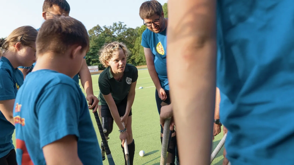 Eine Gruppe Jungendlicher steht auf einem Hockeyplatz. Ein Mädchen mit blonden Haaren scheint den anderen etwas zu erklären.