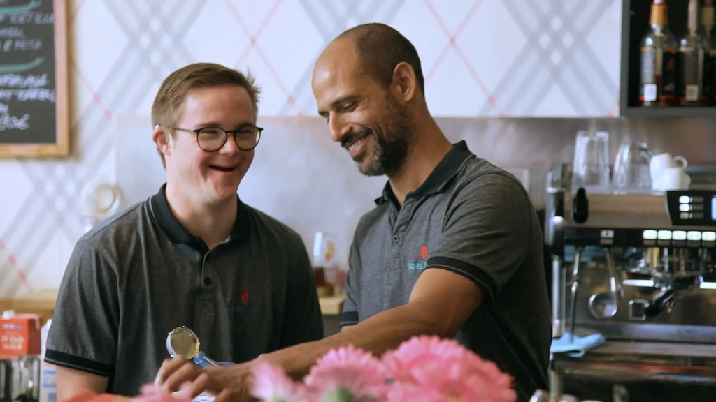 Zwei Männer stehen hinter dem Tresen eins Cafés und lachen. Einer der beiden hat das Downsyndrom.