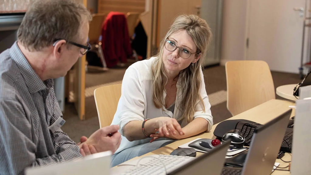Eine Frau und ein Mann sitzen an einem Schreibtisch. Vor ihnen stehen mehrere Laptops, sowie barrierefreie Hilfsmittel zur Bedienung der Laptops. Die Frau erklärt dem Mann die Nutzung eines Hilfsmittels.