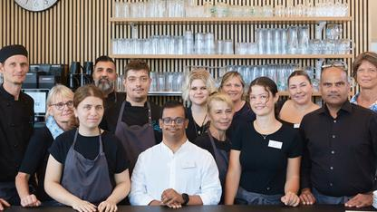Eine Gruppe von Menschen mit und ohne Behinderung steht, mit Schürzen und Küchenkleidung bekleidet, hinter der Theke eines Cafés.