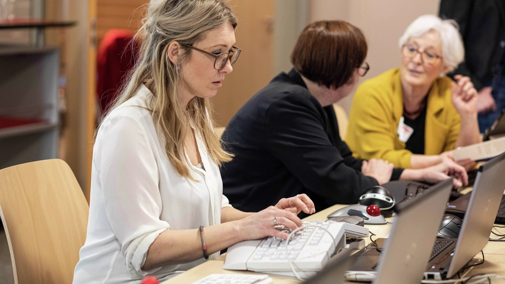 Eine Frau mit langen blonden Haaren und Brille sitzt vor einem Laptop und bedient diesen mit einer ergonomischen Tastatur. Im Hintergrund unterhalten sich zwei Frauen.