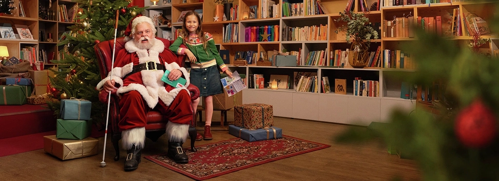 Ein Mann in rot-weißem Weihnachtsmannkostüm sitzt in einem roten Ohrensessel. Im Hintergrund steht ein geschmückter Tannenbaum, drumherum sind Geschenke verteilt. Der Weihnachtsmann hält in seiner rechten Hand einen Blindenstock, in der linken ein Glücks-Los der Aktion Mensch. Neben ihm steht ein junges Mädchen und hält einen Wunschzettel in der Hand. Beide schauen freundlich zur Kamera und lächeln.