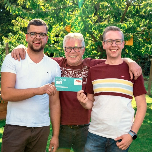 Drei Männer stehen nebeneinander in einem Garten  mit Obstbäumen. Der ältere Mann in der Mitte legt seine Arme um die Schultern der beiden anderen. Die beiden Jüngeren halten gemeinsam ein  Glückslos der Aktion Mensch in den Händen. Alle lächeln glücklich in die Kamera.
