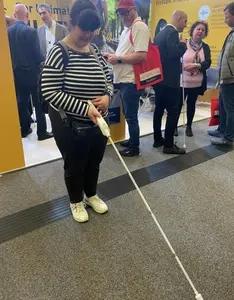 Junge Frau mit dunklen Haaren in gestreiftem Pulli hält an einem Messestand einen Blindenstock in der Hand, der einen verbreiterten Multifunktionsgriff hat, ähnlich einer Fernbedienung.