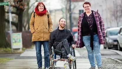Ein Mann im Rollstuhl und zwei Frauen stehen nebeneinander auf dem Bürgersteig.