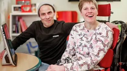 Eine junge Frau und ein junger Mann sitzen nebeneinander vor einem Computer. Die Frau sitzt im Rollstuhl.