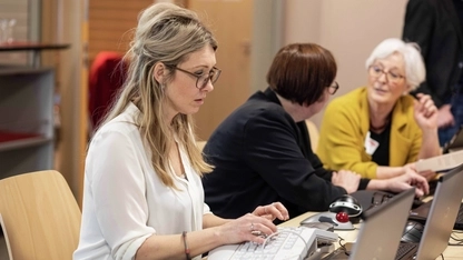 Eine Frau mit langen blonden Haaren und Brille sitzt vor einem Laptop und bedient diesen mit einer ergonomischen Tastatur. Im Hintergrund unterhalten sich zwei Frauen.