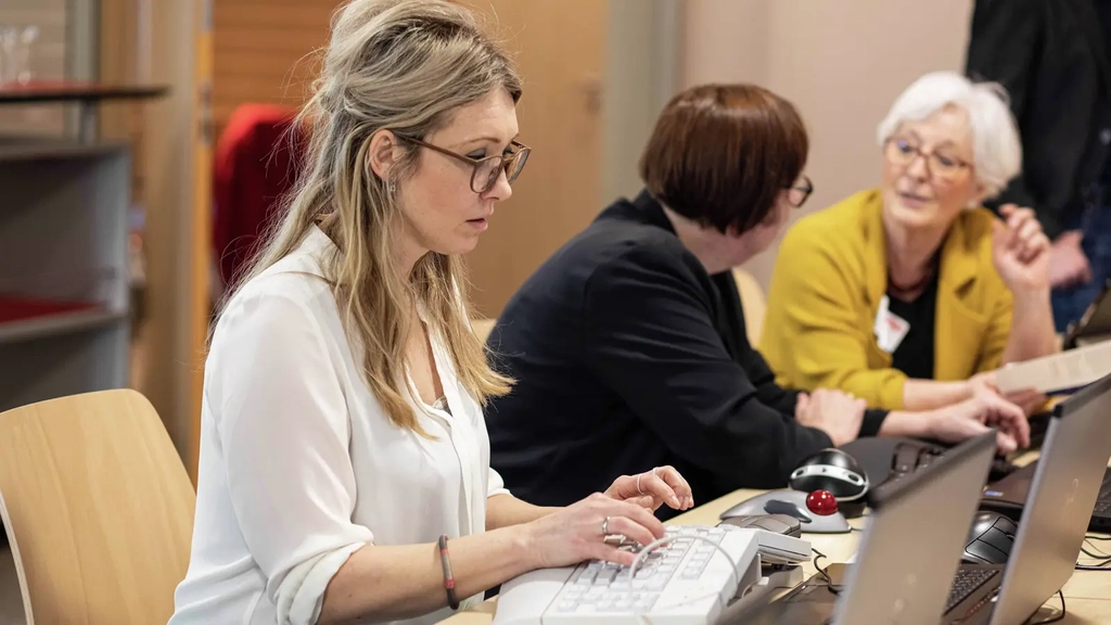 Eine Frau mit langen blonden Haaren und Brille sitzt vor einem Laptop und bedient diesen mit einer ergonomischen Tastatur. Im Hintergrund unterhalten sich zwei Frauen.