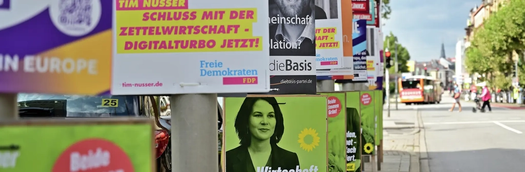 Im rechten Bildrand ist eine Straße zu sehen. Auf dem Bürgersteig auf der linken Bildhälfte sind viele, bunte Wahlplakate an Laternenpfählen angebracht.