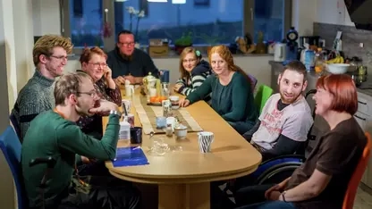 Menschen mit und ohne Behinderung sitzen an einem Tisch und lächeln in die Kamera.