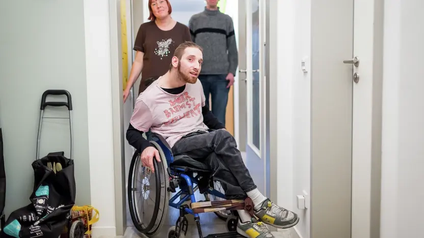 Ein Mann im Rollstuhl fährt durch eine Tür von einem Flur in ein Zimmer. Hinter ihm stehen eine Frau und ein weiterer Mann.