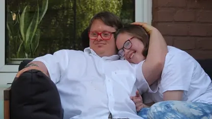 Ein junges Pärchen - beide mit Downsyndrom - sitzen auf dem Sofa. Er hat seinen Arm um sie gelegt und sie kuschelt sich an ihn an.