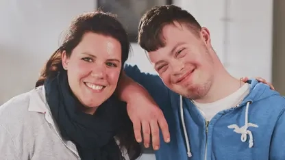 Eine Frau mit braunen Haaren und ein junger Mann mit Down-Syndrom haben einen Arm umeinander gelegt und lachen in die Kamera.