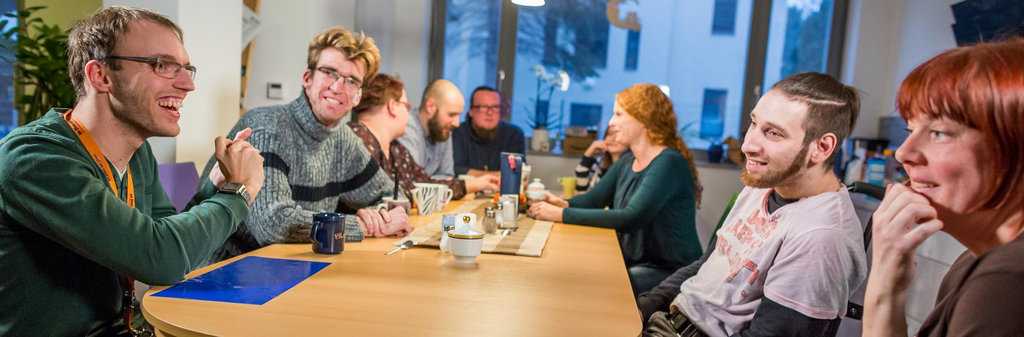 An einem Esstisch sitzen viele verschiedene Menschen mit und ohne Behinderung.