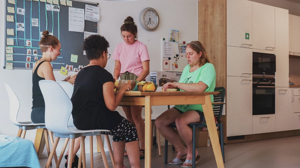 An einem Esstisch in einer Wohnküche sitzen vier Personen und schneiden Gemüse.