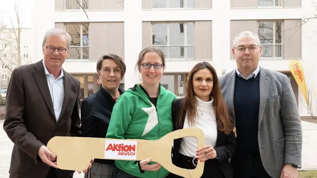 Vertreter*innen der Aktion Mensch, der Stephanus-Stiftung und neue Bewohnerinnen des inklusiven Wohnprojekts der Aktion Mensch steht vor dem Haus in Berlin und halten alle gemeinsam einen großen goldenen Schlüssel in der Hand, auf dem das Aktion Mensch Logo zu sehen ist.