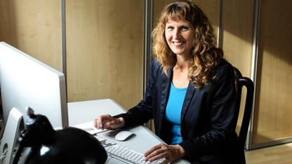 Eine Frau mittleren Alters mit langen dunkelblonden Locken sitzt an einem Schreibtisch. Vor ihr stehen eine Computermaus, eine Tastatur und ein Bildschirm. Sie trägt ein hellblaues T-Shirt und einen dunkelblauen Blazer.