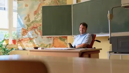 Ein junger Mann in einem Elektrorollstuhl sitzt hinter einem Lehrerpult in einem Klassenzimmer. Hinter ihm sind eine große Landkarte und die Tafel zu sehen.