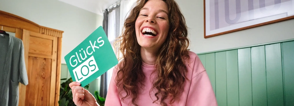 Eine Frau mit braun gelockten langen Haaren sitzt im Schlafanzug auf ihrem Bett. Sie strahlt in die Kamera. In der Hand hält sie das grüne Glücks-Los der Aktion Mensch.