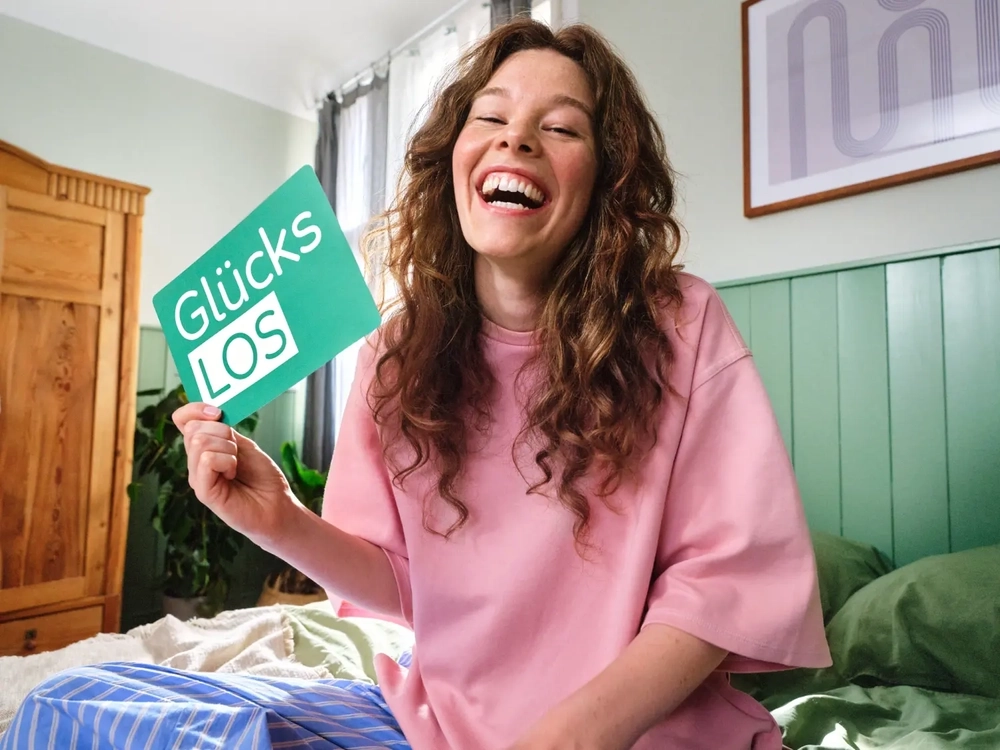 Eine Frau mit braun gelockten langen Haaren sitzt im Schlafanzug auf ihrem Bett. Sie strahlt in die Kamera. In der Hand hält sie das grüne Glücks-Los der Aktion Mensch.