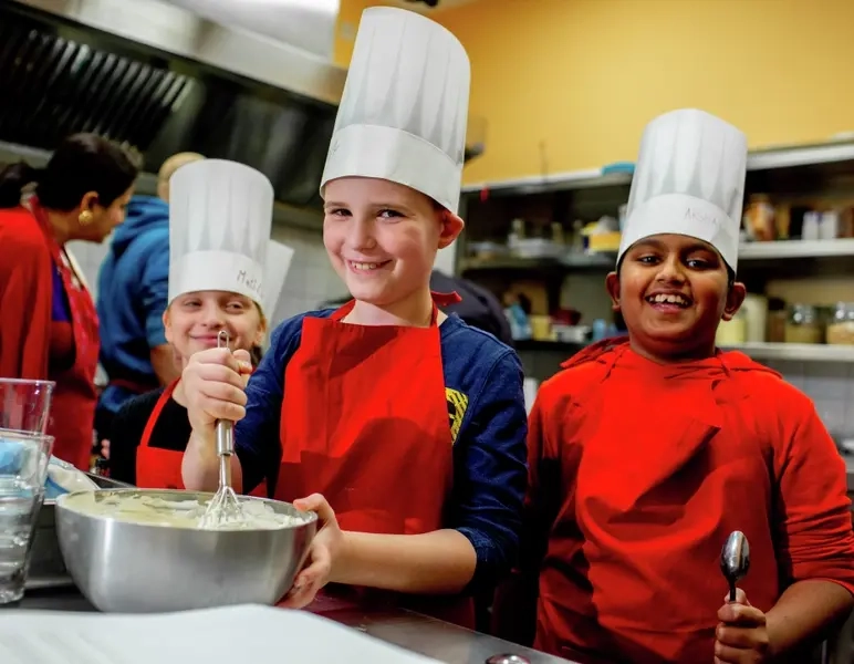 Drei Kinder mit Kochschützen und Kochmützen lachen in die Kamera.