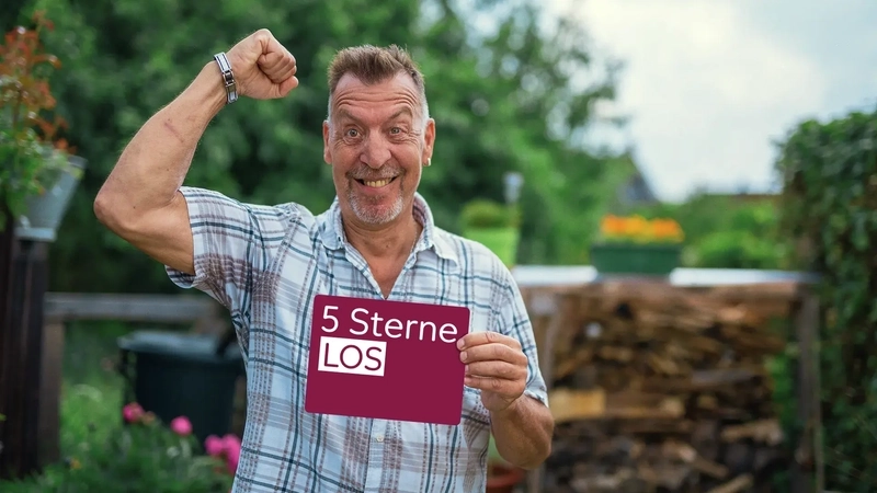 Ein Mann steht im Garten. Er streckt einen Arm jubelnd in die Luft und hält mit der anderen Hand ein 5-Sterne-Los.