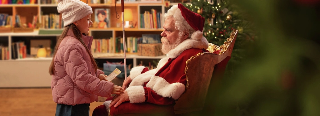 Ein Mann in rot-weißem Weihnachtsmannkostüm sitzt in einem Ohrensessel. Im Hintergrund steht ein geschmückter Tannenbaum. Der Weihnachtsmann hält in seiner rechten Hand einen Blindenstock. Vor ihm steht ein junges Mädchen und hält einen Wunschzettel in der Hand. Sie schaut den Weihnachtsmann freundlich an und legt ihre rechte auf seine linke Hand.