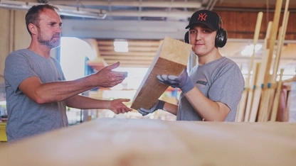 Zwei Männer in einer Holzverarbeitungswerkstatt. Der jüngere der beiden trägt Ohrenschutz und Handschuhe. Er hat einen großen Holzbalken in den Händen. Diesen reicht er in Richtung des anderen Mannes.