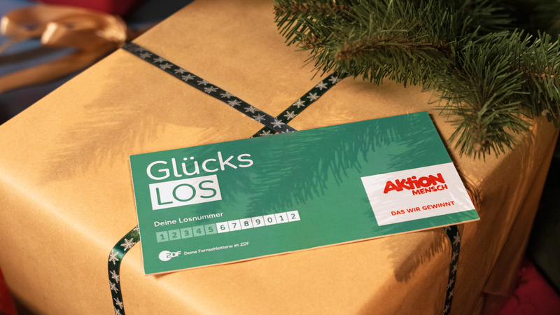Unter einem Weihnachtsbaum liegt ein Geschenk. Auf diesem Geschenk liegt ein Glücks-Los der Aktion Mensch.