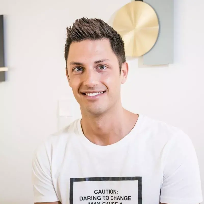 Ein junger Mann mit kurzen, dunklen Haaren. Er trägt ein weißes T-Shirt und lächelt in die Kamera.