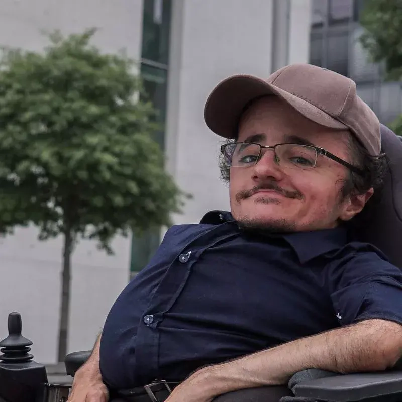 Ein junger Mann mit dunkelblauem Hemd und brauner Kappe. Er sitzt im Rollstuhl und lächelt in die Kamera.