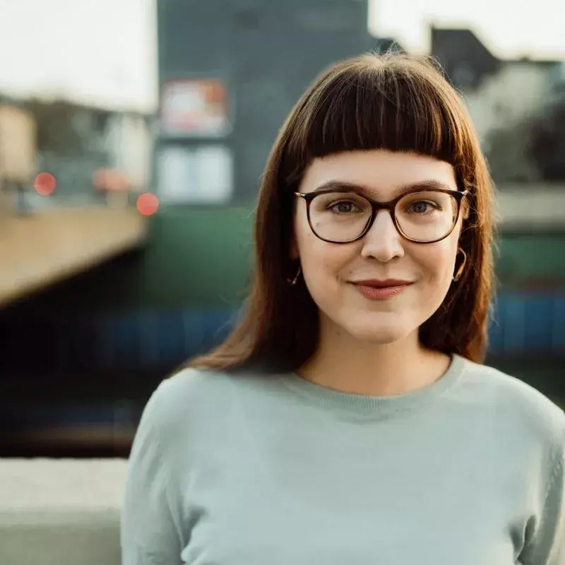 Eine junge Frau mit schulterlangem Haar schaut freundlich in die Kamera. Sie trägt eine Brille und einen blassgrünen Pullover.