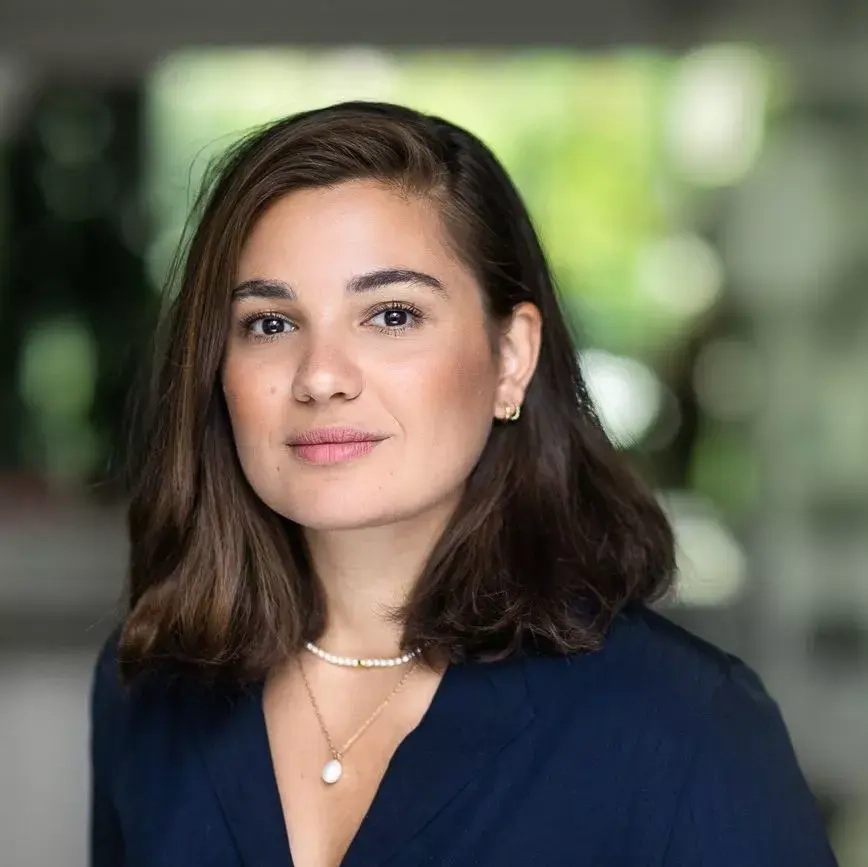 Eine junge Frau mit mittellangen braunen Haaren und einer eichten, blauen Bluse schaut in die Kamera.