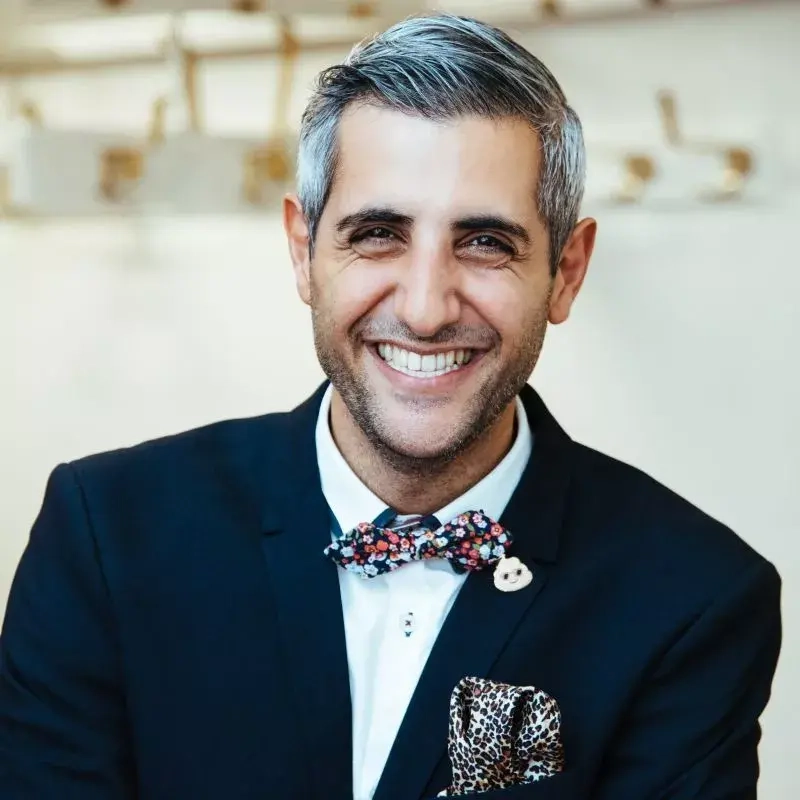 Ein junger Mann mit leicht grauem Haar lächelt freundlich in die Kamera. Er trägt ein dunkelblaues Sakko und eine bunte Fliege.