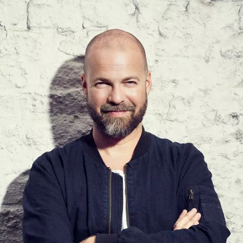 Ein Mann mit Halbglatze und Bart steht vor einer hellen Backsteinwand und blickt freundlich in die Kamera.