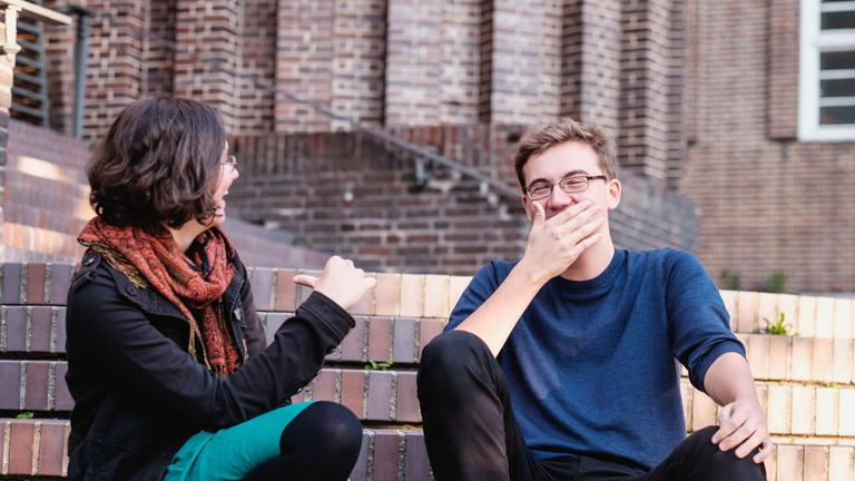 Carlotta - ein Frau mit halblangen brauen Haaren und Brille - und Jasper - ein schlanker Mann mit kurzen dunkelblonenden Haaren und Brille - sitzen auf Treppenstufen und lachen miteinander.