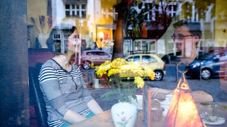 Blick durch das Fenster eines Cafés: dahinter sitzen Carlotta und Jasper an einem Tisch.