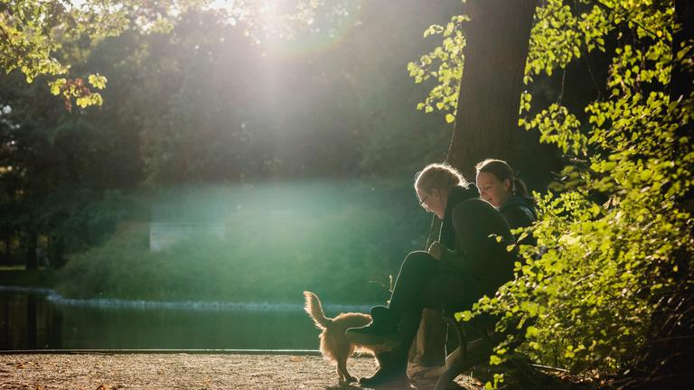 Frauke und Ulrike sitzen mit Hund am See in der Sonne.