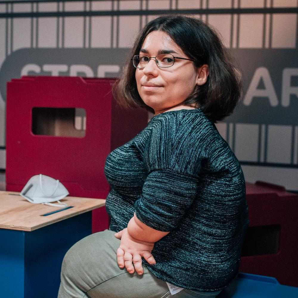 Eine junge, kleinwüchsige Frau sitzt in einem Studio auf einem Hocker 