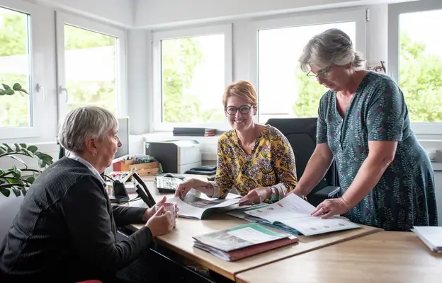 Annette Hambach-Spiegler sitzt am Schreibtisch, sie bespricht mit zwei anderen Frauen eine Broschüre, beziehungsweise sie sehen sich die Broschüre an